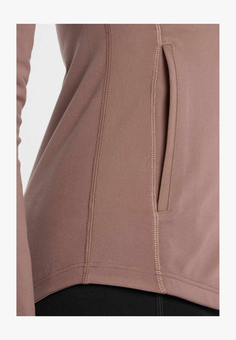 Top atlético de color malva claro hecho de tejido elástico, que cuenta con un bolsillo lateral, costuras planas y un dobladillo curved para facilitar el movimiento.