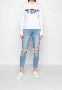 Sudadera blanca con letras en azul marino, combinado con jeans claros rasgados. Zapatillas blancas completan el conjunto casual.