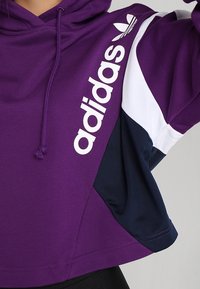 Felpa con cappuccio viola e accenti bianchi e blu navy, con il logo "adidas" bianco presente sul lato sinistro. Tessuto in misto cotone, design cropped.