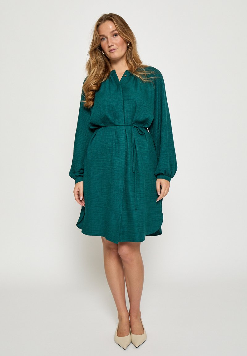 Obleka z ovratnikom v barvi modrozelen kroj z dolgimi rokavi, izdelana iz teksturirane tkanine z rahlo razširjenim spodnjim delom. Oblečena z bež petami.