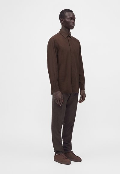 Homme debout portant une chemise marron à manches longues boutonnée, un pantalon marron foncé et des baskets marron sur un fond blanc uni.