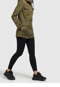 Olive green parka with multiple front pockets, a drawstring waist, and a hood, paired with black ribbed leggings and black sneakers.