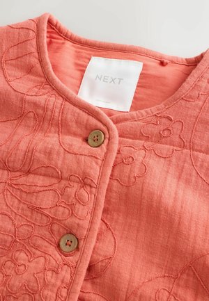 Haut en tissu matelassé rose corail avec broderie florale, encolure ronde, deux boutons en bois, et une étiquette blanche NEXT à l'intérieur du col.
