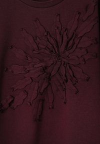 Mørk bordeauxrød stof med et hævet, tekstureret blomsterdesign lavet af lagdelte, syede former, der stråler ud fra midten.