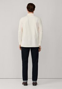 Polo a maniche lunghe crema con colletto, abbinato a jeans blu scuro e scarpe marroni, visto da dietro. Design semplice e pulito.