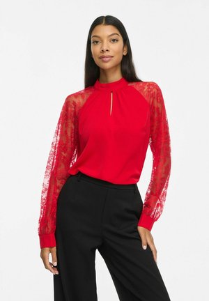 Un mannequin pose sur un fond blanc, portant un chemisier rouge à manches en dentelle transparente et un pantalon noir.