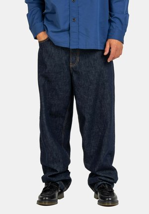 Dunkelblaue Denim-Breite-Hosen mit orangefarbenen Nähten, Fronttaschen, getragen mit einem blauen Hemd und schwarzen Schuhen.
