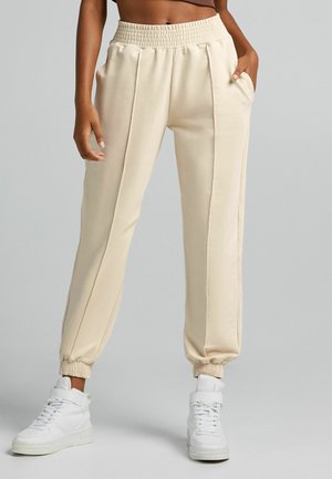 Osoba w beżowych spodniach dresowych z elastycznym pasem i mankietami, w połączeniu z białymi butami sportowymi za kostkę, stojąca na jednolitym tle.