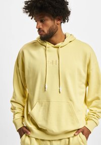 Manteau à capuche jaune en matériau doux, doté d'une poche avant, de cordons avec des embouts blancs et d'un logo en relief discret sur la poitrine.