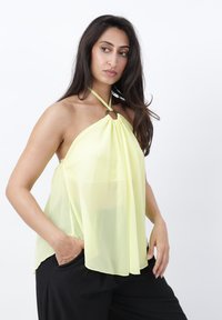 Mujer con el pelo largo y oscuro lleva un top halter amarillo transparente y pantalones negros, posando con una mano en el bolsillo contra un fondo blanco.