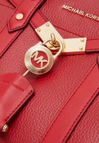 Sac en cuir rouge avec une surface texturée, des finitions en métal doré et un pendentif logo circulaire avec "MK". Gravure visible sur une étiquette en cuir.