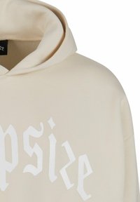 Cremfarbener Kapuzenpullover mit einem großen weißen grafischen Textdesign auf der Vorderseite. Der Stoff wirkt weich und hat eine geräumige Kapuze.