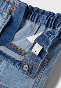 Pantaloni in denim blu con vita elastica, patta con zip e dettagli in rivetti di rame. Presentano cuciture a bordo sfrangiato e un'etichetta bianca all'interno.