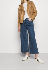Veste de moto en cuir tan, haut texturé blanc, jeans en denim bleu à larges jambes, et baskets blanches avec des accents noirs.