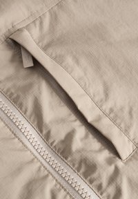 Bežová, textúrovaná tkanina s viditeľným zipsom a malým otvorom na vrecko. Obsahuje mriežkový vzor a prešité detaily pozdĺž okrajov.