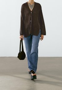 Person wearing a dark brown buttoned cardigan, blue jeans, platform sandals, and holding a small black handbag.
