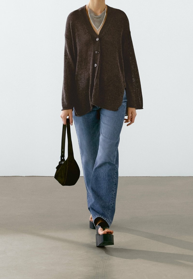 Person wearing a dark brown buttoned cardigan, blue jeans, platform sandals, and holding a small black handbag.