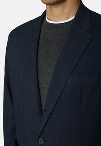 Marinblazer i texturerad tyg, med enkel knapplåsning, utsvängda reverser och bröstficka, bärs över en mörkgrå tröja.