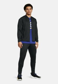 Black zip-up bomber jacket over a blue shirt with yellow accents, paired with black joggers and black athletic shoes with blue laces.