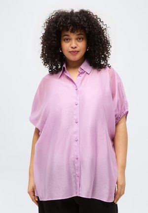 Woman with curly hair wearing a loose, short-sleeved, light purple button-up shirt standing against a plain light background.