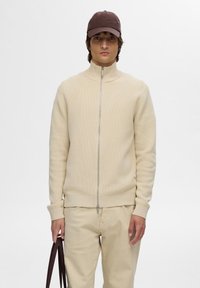 Beige Zip-up-Pullover mit hohem Kragen und strukturiertem Strick, kombiniert mit hellbeigen Hosen und einer dunkelbraunen Mütze. Hält eine braune Tragetasche.
