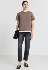 Camicia marrone a maniche corte con uno strato bianco sotto, abbinata a jeans neri tapered e scarpe slip-on nere con dettagli circolari.
