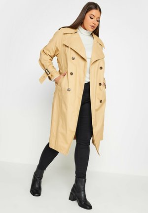 Beige trenchcoat met een dubbelknoopdesign, grote revers en gespsaccenten op de mouwen, gecombineerd met zwarte broek en enkellaarsjes.