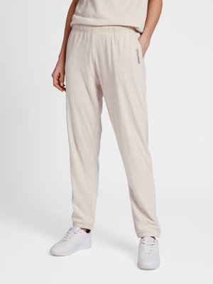 Osoba ubrana w beżowe casualowe spodnie jogger z elastycznym pasem i ściągaczami, w połączeniu z białymi trampkami, stoi z jedną ręką w kieszeni.