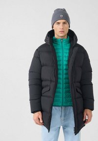 Hombre con un gorro gris, chaqueta acolchada verde azulado, abrigo acolchado negro y jeans de dos tonos azules contra un fondo claro y sencillo.