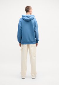 Felpa con cappuccio blu chiaro dal fit rilassato, abbinata a pantaloni cargo color crema con dettagli sulle tasche. Scarpe da ginnastica bianche completano il look.