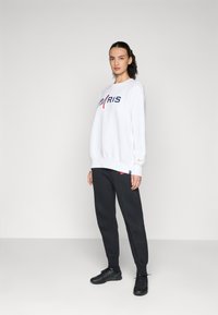 Hvit sweatshirt med "PARIS" i marineblå og røde bokstaver, kombinert med mørkegrå joggebukser og svarte treningssko. Glatt stoff, avslappet passform.