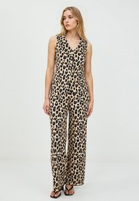 Vrouw in een mouwloze, geknoopte top met luipaardprint en bijpassende wijde broek, met zwarte sandalen, staand tegen een effen witte achtergrond.