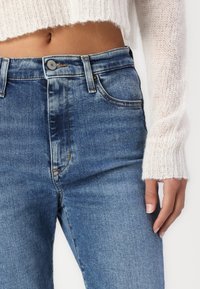 Jeans de mezclilla azules de cintura alta con un ligero desvanecimiento, cierre de botón y dos bolsillos delanteros, combinados con un suéter de punto blanco texturizado.