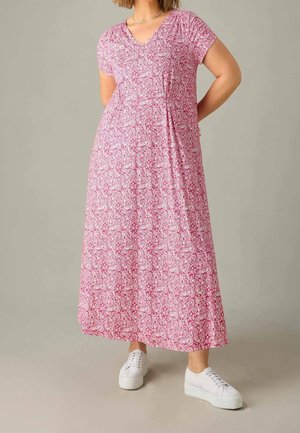 Frau trägt ein langes Maxikleid mit kurzen Ärmeln in pink-weißem Blumenmuster und weiße Plateausneaker, steht mit Händen hinter dem Rücken.