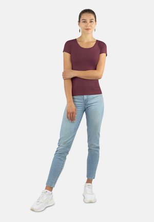 Burgunderfarbendes, tailliertes T-Shirt mit kurzen Ärmeln, kombiniert mit hellblauen Skinny-Jeans und weißen Sportschuhen. Modell steht vor einem neutralen Hintergrund.