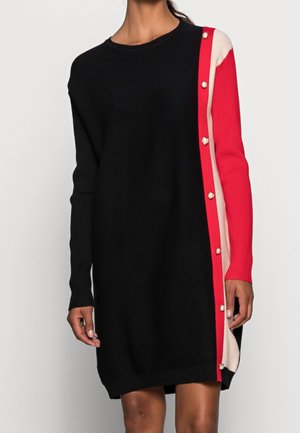 Neulemekko, jossa pitkät hihat, musta etuosan, punainen ja beige värilohkoinen yksityiskohta toisella puolella sekä helminappikoristus punaisen paneelin kohdalla.