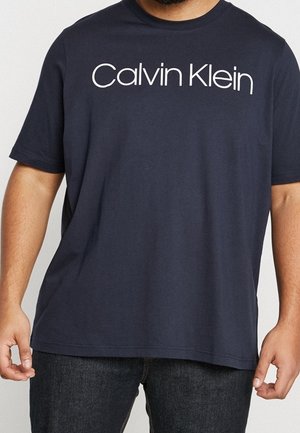 Mężczyzna ubrany w granatowy t-shirt Calvin Klein i ciemne dżinsy, stoi z ramionami luźno opuszczonymi wzdłuż ciała na jednolitym tle.