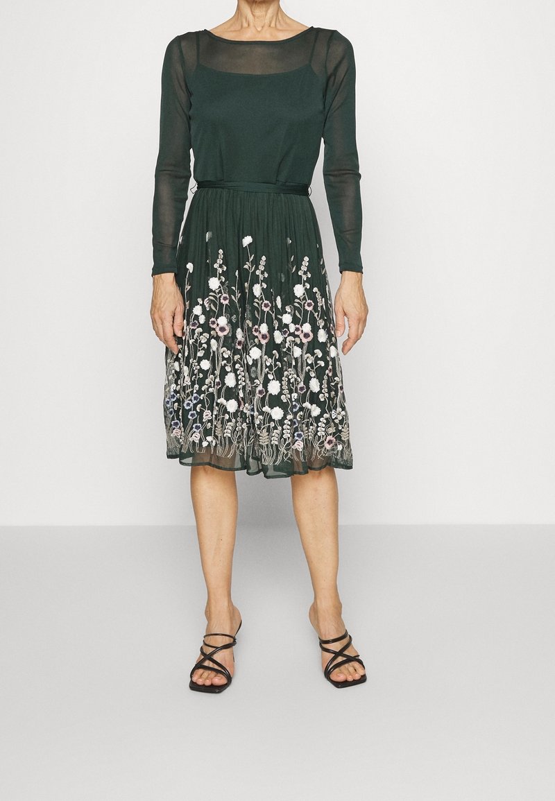 Dunkelgrünes Kleid mit einem transparenten Oberteil mit langen Ärmeln und gesticktem Blumendesign am Saum. Kombiniert mit schwarzen Riemchensandalen.