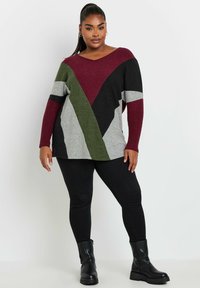 Maglione oversized a V in grigio, verde, bordeaux e nero con blocchi di colore geometrici. Abbinato a jeans skinny neri e stivaletti.