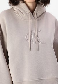 Sudadera de color beige claro con un bolsillo tipo canguro, capucha con cordón y logo bordado "CK" en el pecho. Material suave y corte relajado.