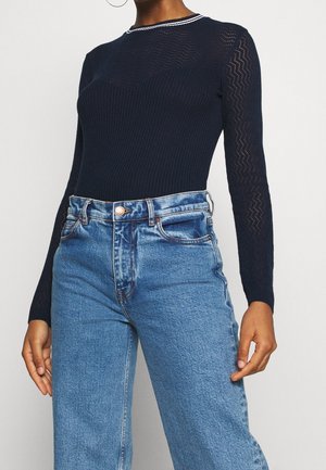 Femme portant un haut côtelé manches longues bleu marine avec des manches à motifs et un jean bleu taille haute sur un fond uni.