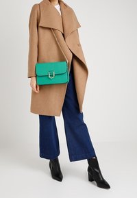 Tansfärgad ullkappa med breda slag, tealfärgad texturerad handväska med lås, och svarta spetsiga högklackade stövlar, kombinerat med blå vida jeans.