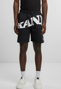 Black shorts with a large white textured "KAND" logo, featuring an elastic waistband and drawstring. Paired with white sneakers and socks.