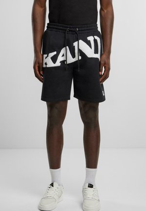 Shorts noirs avec un grand logo texturé blanc "KAND", avec une taille élastique et un cordon de serrage. Associés à des baskets et des chaussettes blanches.