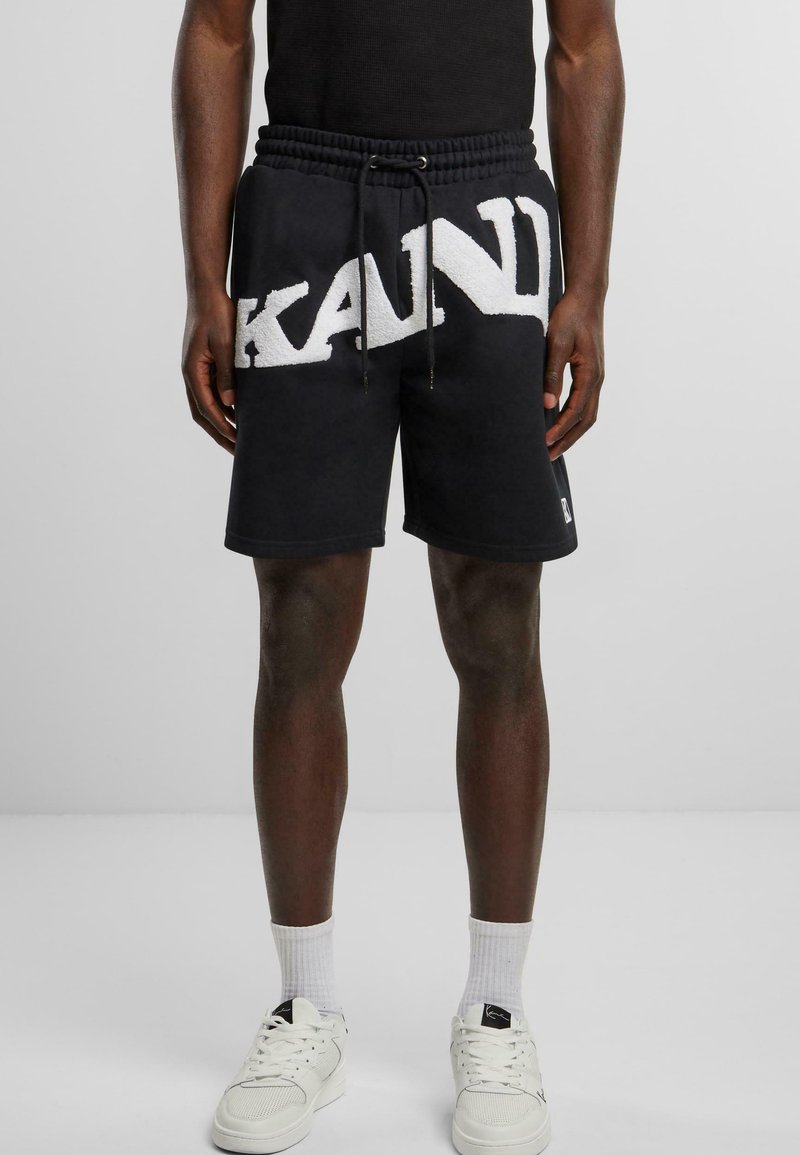 Black shorts with a large white textured "KAND" logo, featuring an elastic waistband and drawstring. Paired with white sneakers and socks.