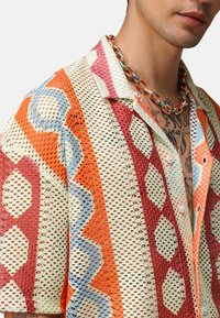 Koszula z krótkim rękawem z koronkowym wzorem z mieszanką geometrycznych motywów w odcieniach czerwieni, pomarańczy, niebieskiego i kremowego, o lekkiej strukturze.