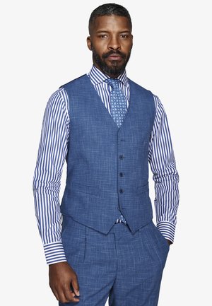 Uomo che indossa un abito blu a tre pezzi con texture, camicia a righe e cravatta con motivo, in piedi con una mano in tasca.