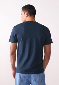 T-shirt bleu marine à manches courtes, en coton, col rond, texture lisse, porté avec un short en denim bleu clair, tatouage visible sur le bras.