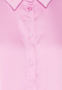 Helles rosa Hemd, Nahaufnahme der Vorderseite mit drei sichtbaren Knöpfen und einem spitzen Kragen auf einer glatten Stoffoberfläche.