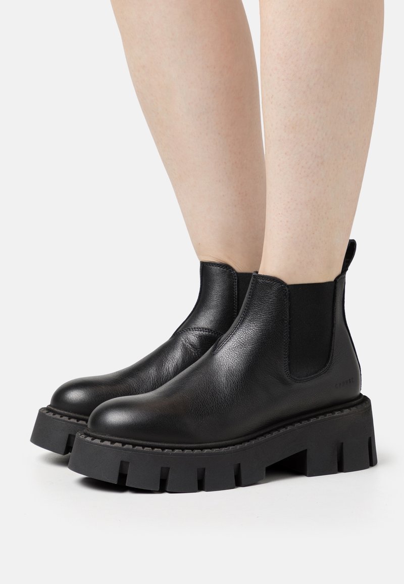 Bottines en cuir noir à semelle épaisse portées sur jambes nues, avec des empiècements latéraux élastiques et un anneau de tirage à l'arrière.